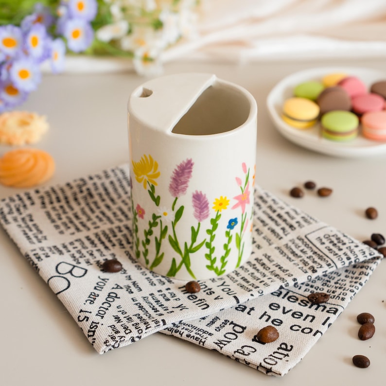 May include: A white ceramic mug decorated with a floral pattern of yellow, purple, and pink flowers. The mug rests on a newspaper-print napkin, with coffee beans scattered around. Macarons are in the background.