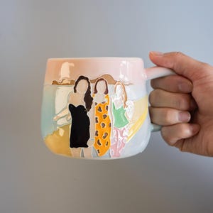 Może przedstawiać: Ceramiczny kubek z wypukłą, kolorową ilustracją trzech postaci na plaży. Postacie mają na sobie czarną sukienkę, pomarańczową sukienkę we wzory oraz zielony top ze wzorzystymi spodniami. Tło przedstawia delikatne niebo i piaszczysty brzeg.