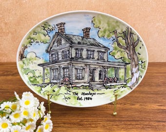 May include: Oval ceramic plate featuring a hand-painted illustration of a two-story house with a porch, trees, and a blue sky. The plate includes the text "The Stanleys - Est. 1984". The plate is displayed on a gold-colored stand.