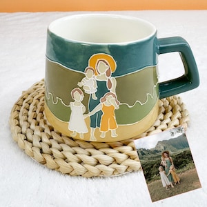May include: A ceramic mug with a green handle and a painted illustration of a mother and children. The mug has a cream-colored interior and sits on a woven coaster. A small photo of a family is next to the mug.