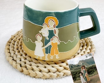 May include: A ceramic mug with a green handle and a painted illustration of a mother and children. The mug has a cream-colored interior and sits on a woven coaster. A small photo of a family is next to the mug.