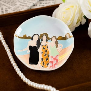 May include: A ceramic dish with a beach scene featuring three figures in dresses. The dish has a light blue sky and a sandy beach design. The figures are in black, orange, and green dresses. A pearl necklace is in the foreground.