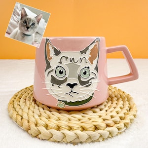 Puede incluir: Una taza de cerámica rosa con un diseño de cara de gato y la palabra "sun" sobre los ojos. La taza tiene un asa cuadrada y se apoya en un posavasos tejido. Una pequeña foto de un gato está en la esquina superior izquierda.