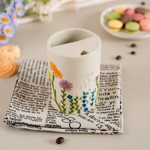 May include: A cream ceramic pen holder decorated with flowers and the text "GOD IS GOOD." It rests on a black and white newspaper print cloth. A plate of macarons is in the background.