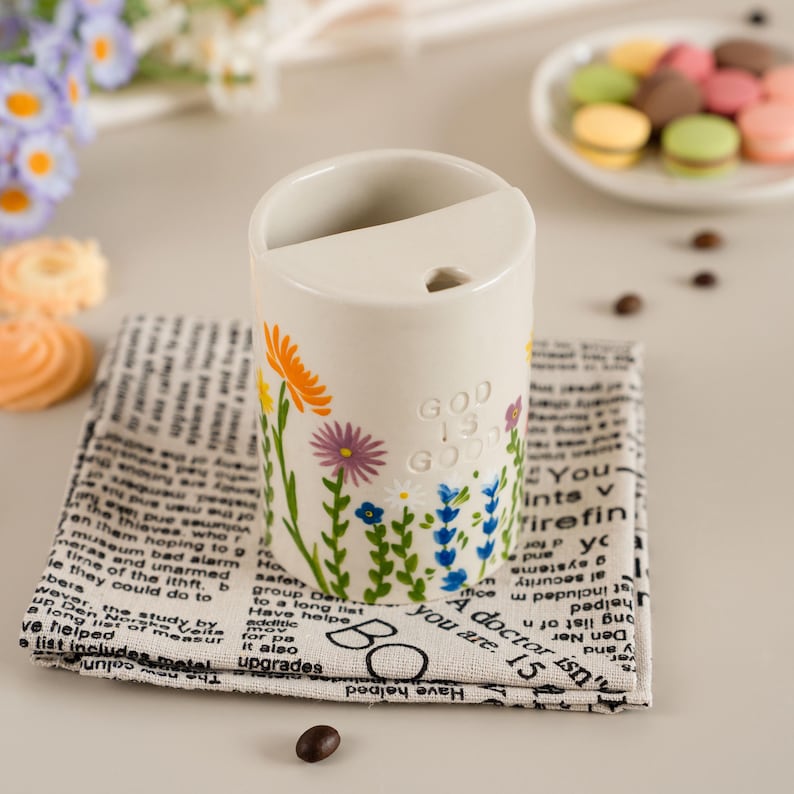 May include: A cream ceramic pen holder decorated with flowers and the text "GOD IS GOOD." It rests on a black and white newspaper print cloth. A plate of macarons is in the background.