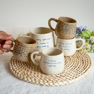 May include: Four ceramic mugs in cream and tan colours, each featuring handwritten blue text. The mugs have rounded bodies and curved handles. The text includes phrases like 'the story isn't over' and 'I love you times infinity to the infinity'.