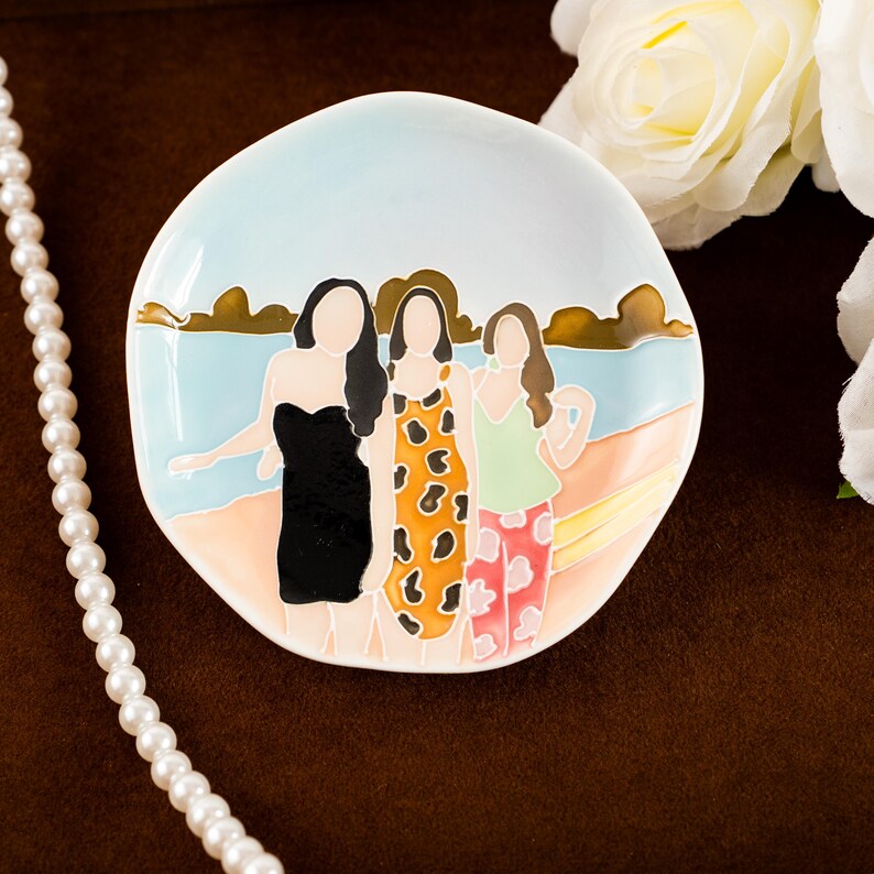 May include: A ceramic dish with a beach scene featuring three figures in dresses. The dish has a light blue sky background with gold accents and a pearl necklace on the side. The figures are in black, orange, and green outfits.