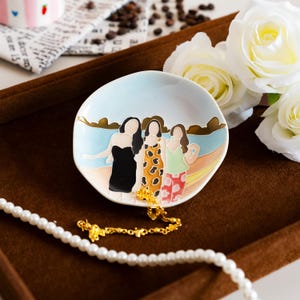 May include: A small, round ceramic dish with a painted beach scene featuring three figures in dresses. The dish has a light blue background and a gold necklace rests on it. White roses and a pearl necklace are in the background.