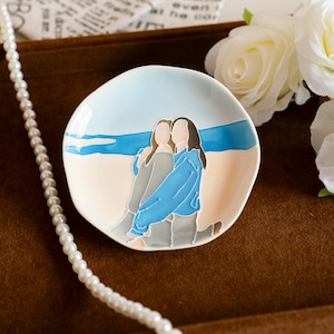 May include: A small, round ceramic dish featuring a beach scene. Two figures are depicted embracing on the beach, with a blue sea and sky in the background. The dish is white with a brown border.