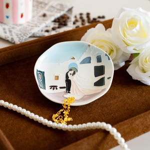 May include: A small, white ceramic dish with a painted scene of a couple kissing in front of a building. The dish has a light blue sky background and is accented with black, blue, and gold details. A gold necklace and a pearl necklace are in the foreground.