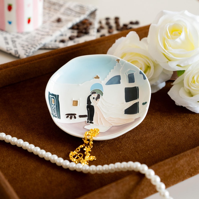 May include: A small, white ceramic dish with a painted scene of a couple kissing in front of a building. The dish has a light blue sky background and is accented with black, blue, and gold details. A gold necklace and a pearl necklace are in the foreground.