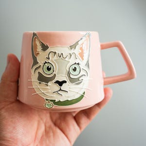 Może przedstawiać: Jasnoróżowy ceramiczny kubek z wypukłą, kolorową ilustracją kociego pyszczka z napisem "sun" nad czołem. Kot ma zielone oczy, szaro-białe znaczenia i zieloną obrożę. Kubek posiada uchwyt o geometrycznym kształcie.