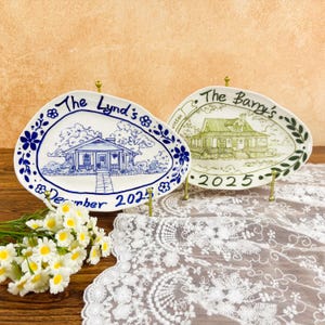May include: Two decorative ceramic plates with house illustrations. One plate is blue and white with the text "The Lynd's" and "December 2025". The other plate is green and white with the text "The Barry's" and "2025". Both plates are on gold stands.