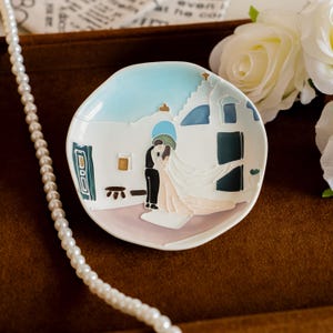 May include: A small, white ceramic dish with a painted scene of a couple embracing in front of a building. The dish has a light blue sky and a pearl necklace is draped nearby. The dish is approximately 10 cm in diameter.