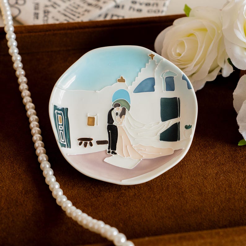May include: A small, white ceramic dish with a painted scene of a couple embracing in front of a building. The dish has a light blue sky and a pearl necklace is draped nearby. The dish is approximately 10 cm in diameter.