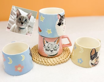 May include: A collection of ceramic mugs with cat and dog designs. The mugs feature various colors, including blue, pink, and yellow, with illustrations of cat faces, stars, moons, and flowers. One mug has a French bulldog design.