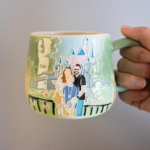 May include: A light green ceramic mug featuring a raised, colourful relief design of a family of three standing in front of a castle. The illustration includes soft pastel tones, a black fence at the base, and a handle extending from the right side.
