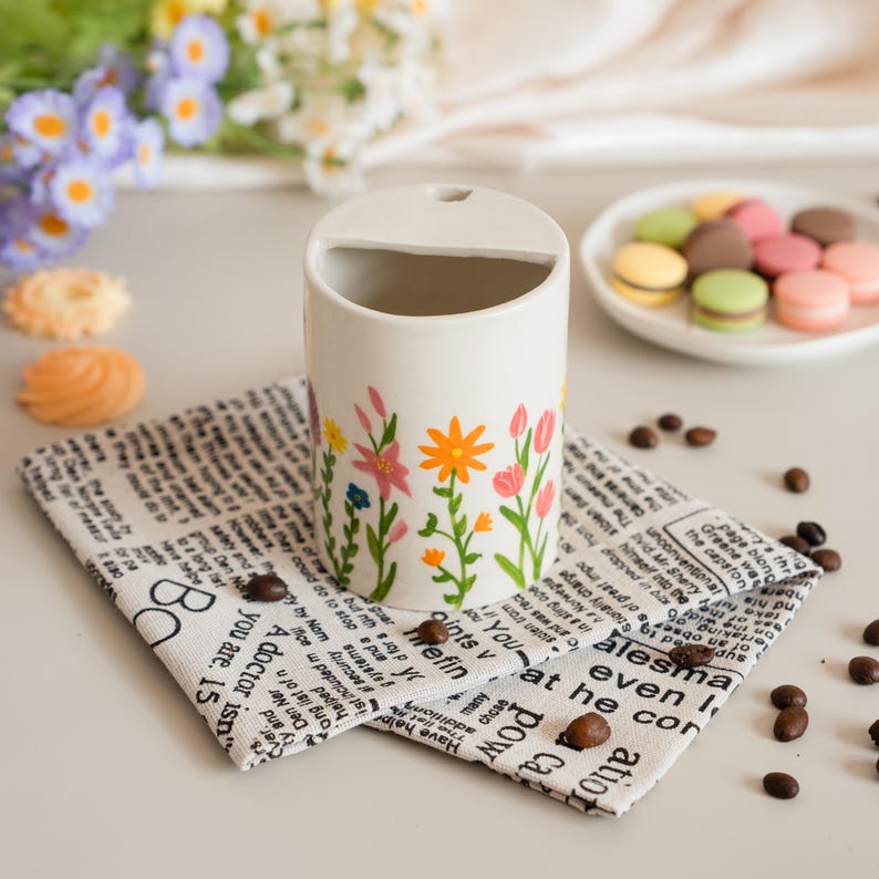 May include: A cream-coloured ceramic utensil holder decorated with a floral pattern in pink, orange, and blue. It rests on a newspaper-print napkin, with coffee beans scattered around. A plate of macarons is in the background.
