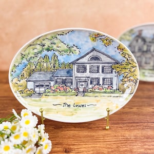 May include: Oval ceramic plate with a hand-painted illustration of a house, trees, and a blue sky. The plate is white with a gold stand and the text "The Lowes" is written below the house.