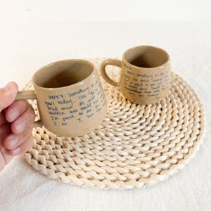 Puede incluir: Dos tazas de cerámica beige con texto azul escrito a mano. Las tazas tienen forma redondeada y un asa. El texto de una taza dice: "Aquí hay algo para secar las lágrimas de hoy... Estoy orgulloso de verte... Te amo". Las tazas están sobre un salvamanteles tejido.
