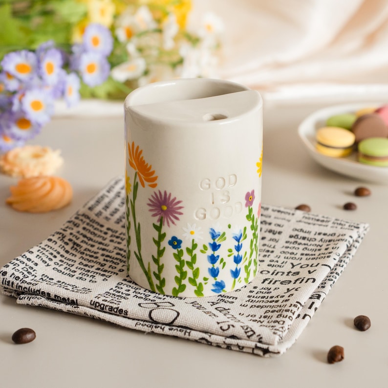 May include: A cream-coloured ceramic container decorated with colourful flowers and the text "GOD IS GOOD." It rests on a newspaper-print cloth. A plate of macarons is in the background.