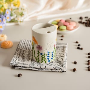 May include: A cream ceramic pen holder decorated with flowers and the text "GOD IS GOOD." It rests on a newspaper-print napkin. Scattered around are colorful flowers and coffee beans. The holder is approximately 10 cm tall.