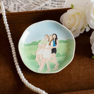 May include: A small, round ceramic dish with a light blue and green landscape design. The dish features an illustration of three figures embracing. A pearl necklace is draped on the side.