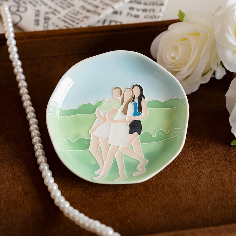 May include: A small, round ceramic dish with a light blue and green landscape design. The dish features an illustration of three figures embracing. A pearl necklace is draped on the side.