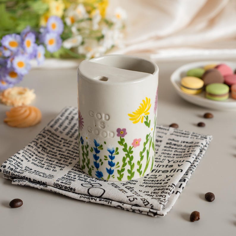 May include: A cream ceramic tumbler decorated with flowers and the text "GOD IS GOOD." It rests on a newspaper-print napkin. A plate of macarons and flowers are in the background.