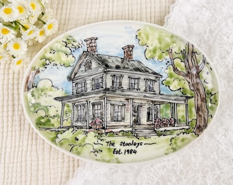 May include: Oval ceramic plate featuring a hand-painted illustration of a two-story house with a porch, trees, and a blue sky background. The plate is inscribed with "The Stanleys - Est. 1984". The color palette includes white, green, brown, and blue.