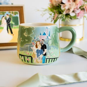 May include: A light green ceramic mug featuring a raised, colorful relief design of a family of three standing in front of a castle. The illustration includes stylized greenery, a black fence, and a pink-topped castle structure on the mug surface.