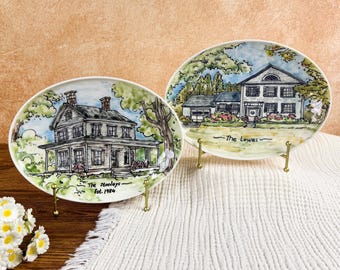 May include: Two decorative plates with hand-painted house illustrations. One plate features a house with the text "The Stanleys Est. 1984". The other plate shows a different house with the text "The Lowes". Both plates are displayed on gold stands.