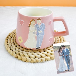 May include: Pink ceramic mug with a square handle, featuring a wedding couple illustration and heart and flower accents. The mug sits on a woven coaster, with a small photo of the couple. The mug is a unique gift idea.
