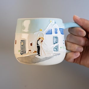 Könnte beinhalten: Ein Keramikbecher mit einem erhabenen, dreidimensionalen Reliefdesign eines Paares in Hochzeitskleidung vor einem weißen Gebäude mit blauen Akzenten. Der Becher hat einen hellblauen oberen Teil, einen weißen Körper und einen geschwungenen Henkel.