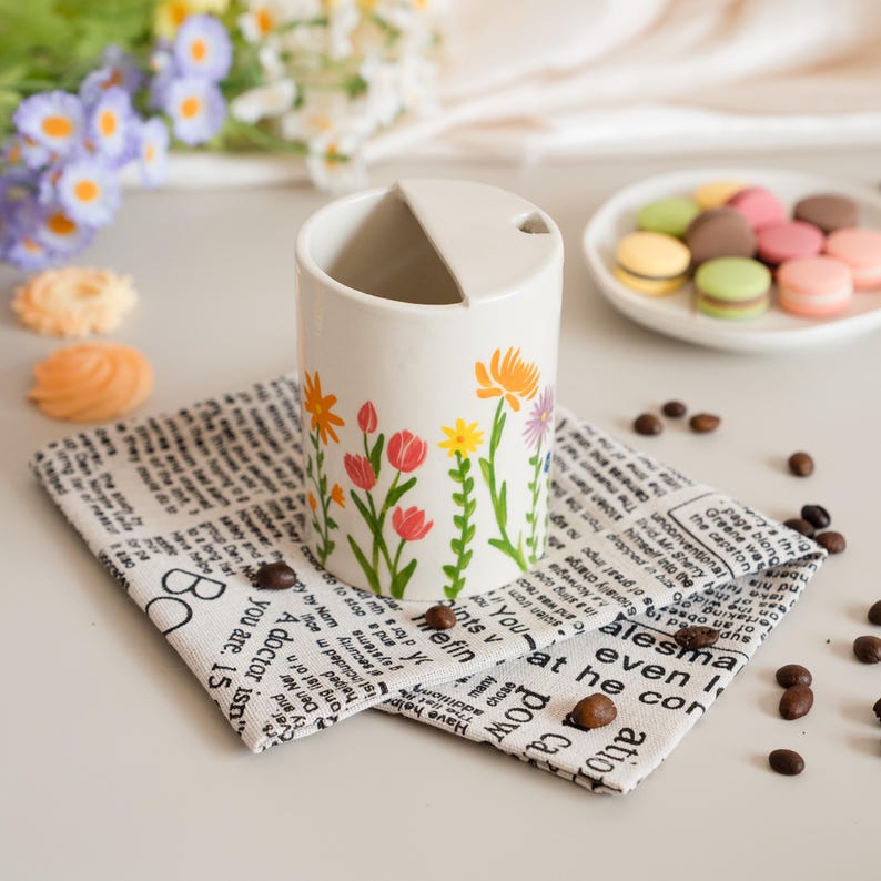 May include: A cream-coloured ceramic container with a lid, decorated with vibrant floral designs. It rests on a newspaper-print cloth, with coffee beans and a plate of macarons nearby. The flowers include red tulips, yellow and orange daisies, and purple flowers.