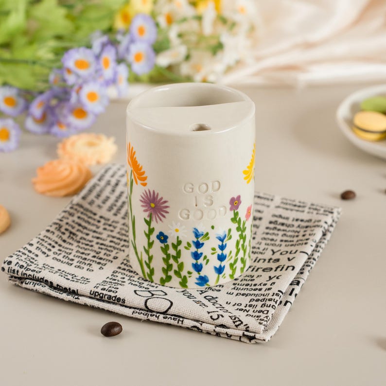 May include: A white ceramic pen holder with a floral design in orange, purple, and blue. The words "GOD IS GOOD" are printed on the side. The pen holder sits on a black and white newspaper print napkin.