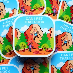 Op de afbeelding: Vierkante sticker met een cartoon illustratie van klimmers op een rode en oranje berg. De sticker heeft de tekst "CAN I PET THAT ROCK" in een blauwe afgeronde rechthoek. Groene bomen en een blauwe lucht met wolken zijn op de achtergrond.