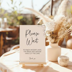 May include: A beige sign with black text that reads "Please Wait" and "To enjoy dessert until the bride and groom have cut the cake. Thank you." The sign is on a wooden easel and is in front of a vase with dried flowers.