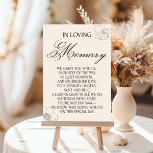 Puede incluir: Un letrero color crema sobre un pequeño caballete de madera, con las palabras "IN LOVING Memory" en una elegante escritura. El letrero incluye un poema sobre el recuerdo y está decorado con ilustraciones florales. Un jarrón de flores secas está en el fondo.