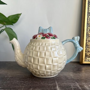 May include: Cream-colored ceramic teapot with a basketweave pattern. The lid is adorned with a cluster of red and white flowers and a light blue bow. The handle and spout have light blue accents, creating a charming, decorative piece.