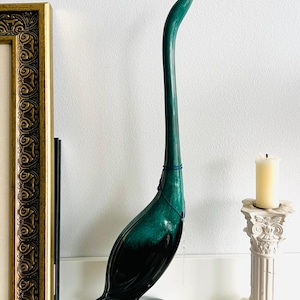 May include: A tall, emerald green ceramic bird sculpture with a long, curved neck and a dark base. The bird is displayed on a white shelf, next to a gold framed picture and a white candle on a decorative stand. The sculpture has a glossy finish.