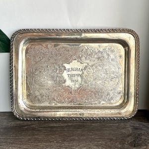 May include: Silver rectangular tray with an ornate floral and scrollwork design. The tray has a rope-like border and features the text "SEAGRAM TROPHY 1951" in the centre. A decorative gold framed object is visible on the right.