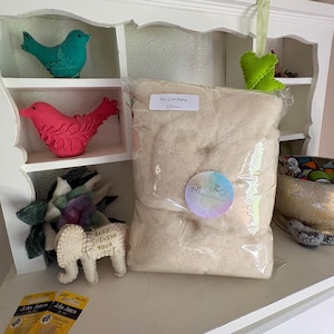 May include: A clear plastic bag filled with beige wool stuffing, labeled "Fine Wool Stuffing". A small, white felt elephant with the text "Keep Shining Your Light" is next to the bag. Decorative felt birds and a bowl are also visible.