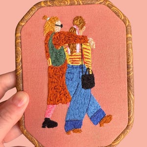 May include: Embroidered artwork depicting two figures, one wearing a green backpack and an orange dress, the other in a yellow and red striped shirt and blue jeans, embracing. The artwork is framed in a wooden frame with a brown and gold pattern.