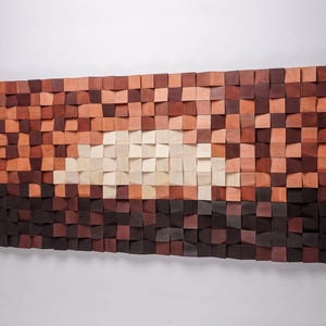 Könnte beinhalten: Ein großes, rechteckiges Wandkunstwerk aus Holz, das aus zahlreichen kleinen, quadratischen Blöcken besteht. Die Blöcke variieren in der Farbe, von Dunkelbraun bis Hellbeige, wodurch ein Gradienteneffekt entsteht. Das Kunstwerk ist an einer weißen Wand befestigt.