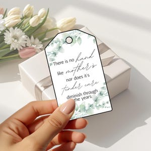 May include: A white gift tag with a floral design and the text "There is no hand like mother's nor does it's tender care diminish through the years." The tag is held in front of a white gift box and flowers.