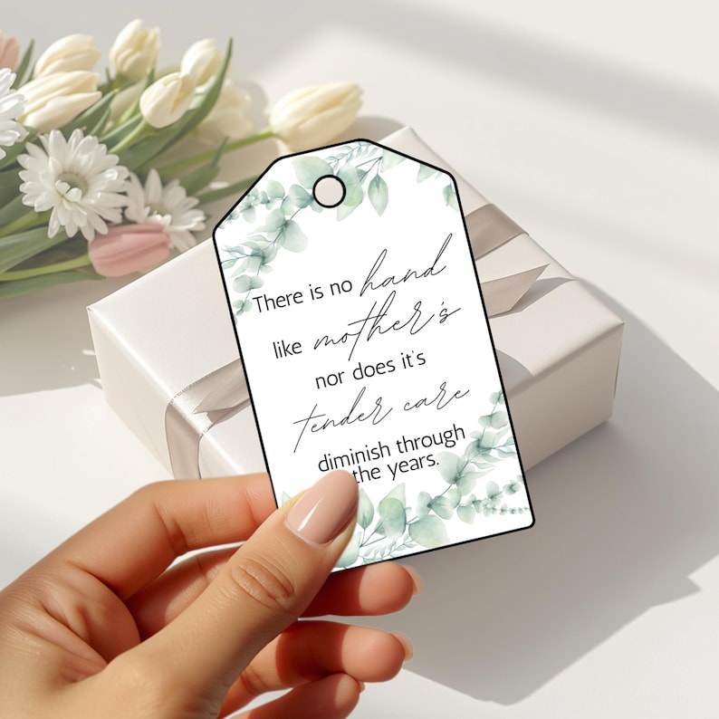 May include: A white gift tag with a floral design and the text "There is no hand like mother's nor does it's tender care diminish through the years." The tag is held in front of a white gift box and flowers.