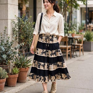 May include: A long, tiered skirt featuring alternating horizontal bands of solid black and a beige floral pattern, paired with a cream-colored button-down shirt with long sleeves and a collar, and light beige low-top sneakers with laces.