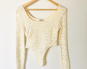 May include: A white lace long-sleeve bodysuit with a scoop neck and a snap closure at the crotch. The bodysuit is hanging on a wooden hanger against a white wall. The brand name "ACDC" is visible on the front of the bodysuit.
