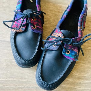 Puede incluir: Un par de zapatos náuticos negros con detalles de tela estampada colorida en la parte superior y alrededor de los tobillos. Los zapatos tienen cordones negros y un diseño cosido alrededor de la puntera.